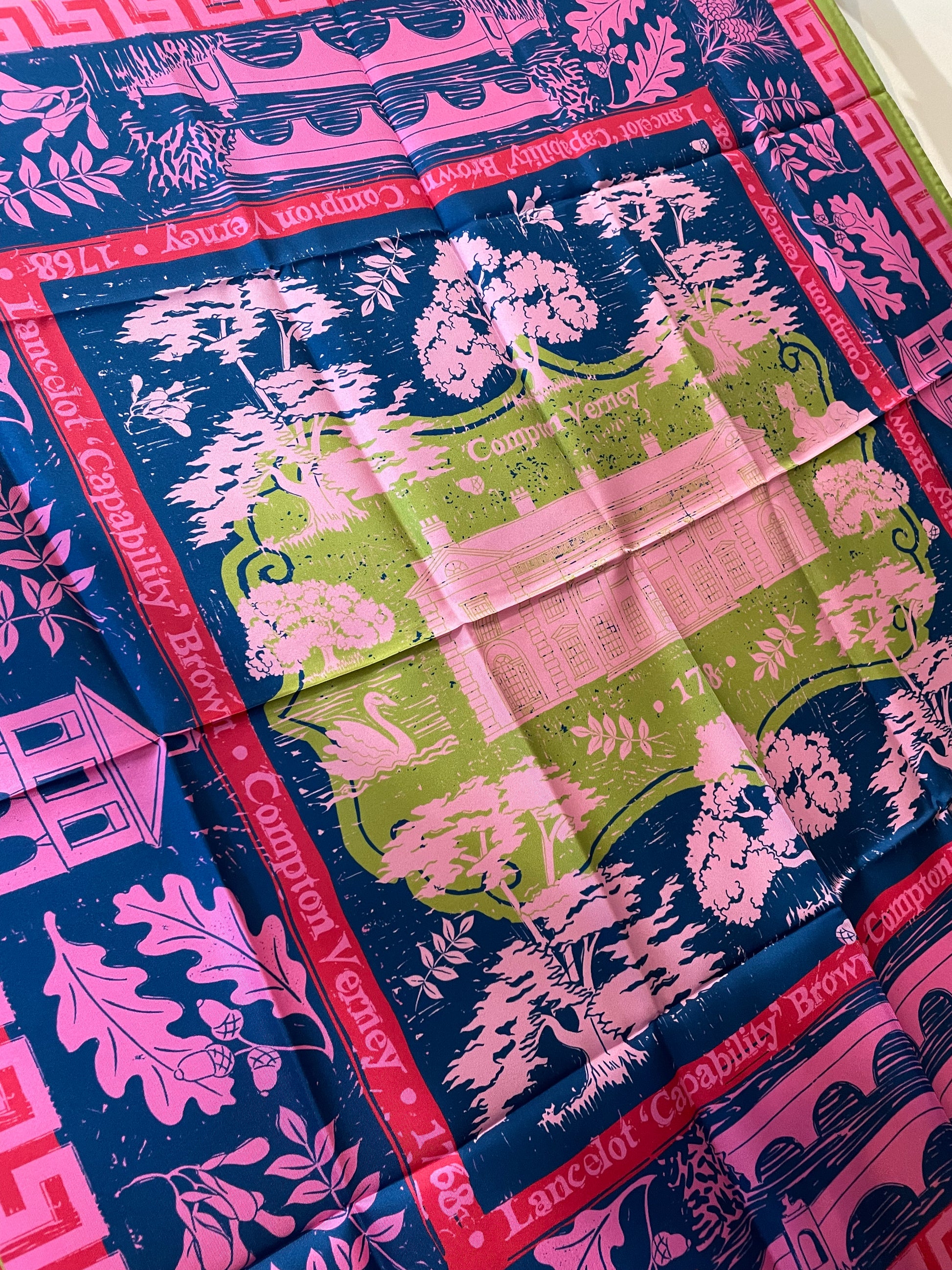 A close up detail of the scarf showing a design of trees, leaves, buildings and nature in pink against a dark blue background with Compton Verney shown in pink on a light green background in the centre.