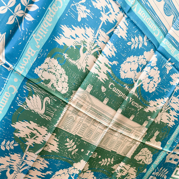 A close up detail of the scarf showing a design of trees, leaves, buildings and nature in light grey against a blue background with Compton Verney shown in light grey on a dark green background in the centre.