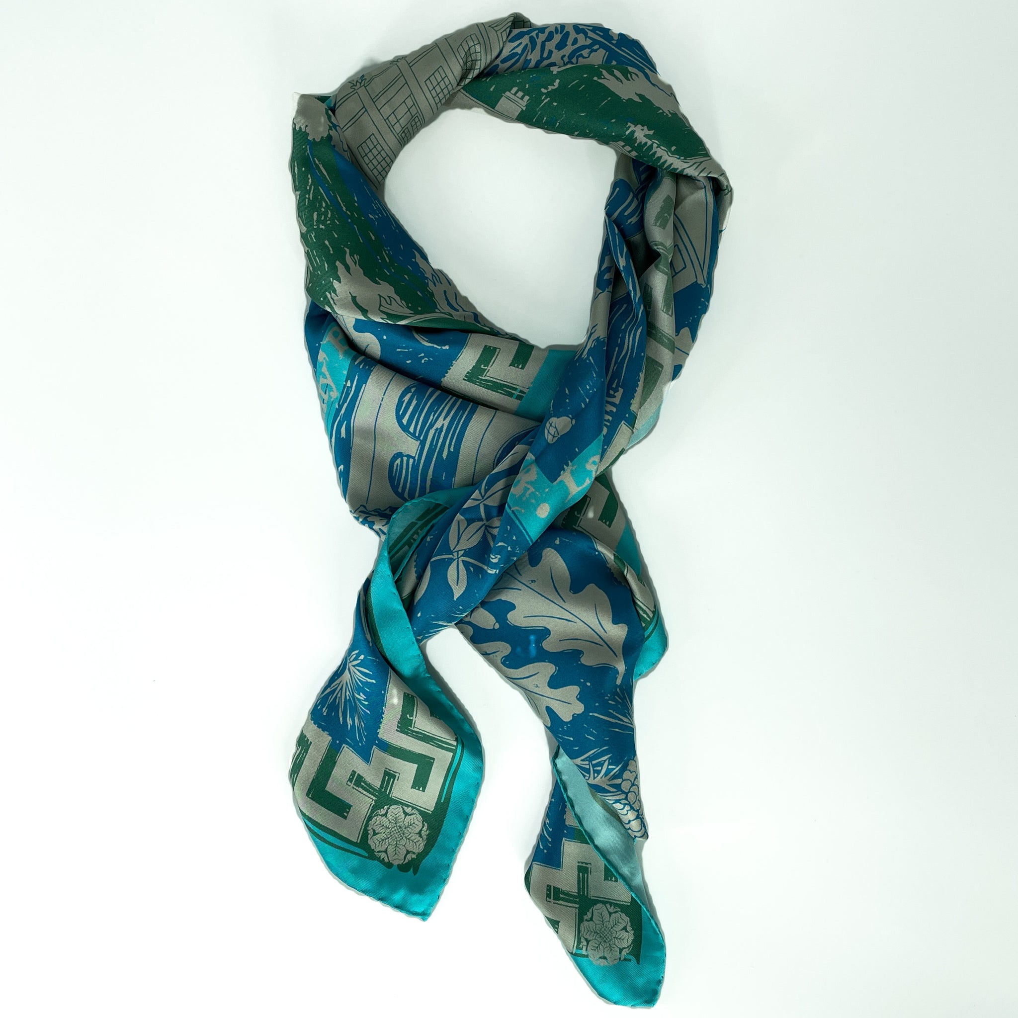 A silk scarf laid out in a knot shape with shades of light blue, dark blue, green and grey from the geometric design.